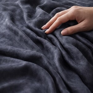 K&ouml;nnte beinhalten: Dunkelblauer Stoff mit weichem, strukturiertem Aussehen. Der Stoff ist drapiert und erzeugt sanfte Falten und Schatten. Eine Hand ber&uuml;hrt sanft den Stoff und hebt seine Textur und seinen Fall hervor. Das Thema ist Textilien.