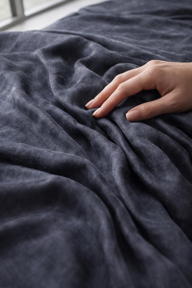 K&ouml;nnte beinhalten: Dunkelblauer Stoff mit weichem, strukturiertem Aussehen. Der Stoff ist drapiert und erzeugt sanfte Falten und Schatten. Eine Hand ber&uuml;hrt sanft den Stoff und hebt seine Textur und seinen Fall hervor. Das Thema ist Textilien.