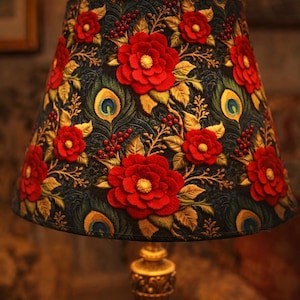 May include: A decorative lampshade featuring a dark background with embroidered red flowers, gold leaves, and peacock feather accents. The lampshade sits atop a gold-colored lamp base, creating a warm, inviting ambiance.