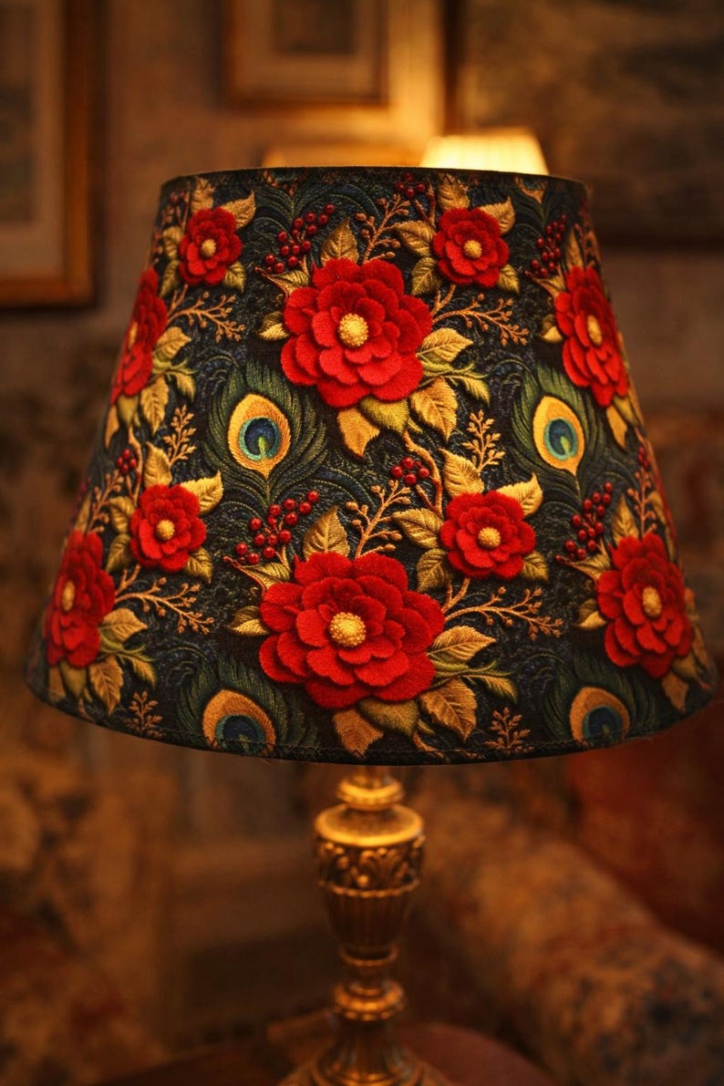 May include: A decorative lampshade featuring a dark background with embroidered red flowers, gold leaves, and peacock feather accents. The lampshade sits atop a gold-colored lamp base, creating a warm, inviting ambiance.
