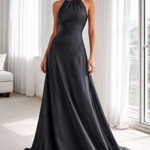 K&ouml;nnte beinhalten: Ein dunkelgraues Neckholder-Kleid mit flie&szlig;endem A-Linien-Rock. Das Kleid hat ein figurbetontes Oberteil und einen bodenlangen Saum. Das Model steht vor einem Fenster mit wei&szlig;en Vorh&auml;ngen.