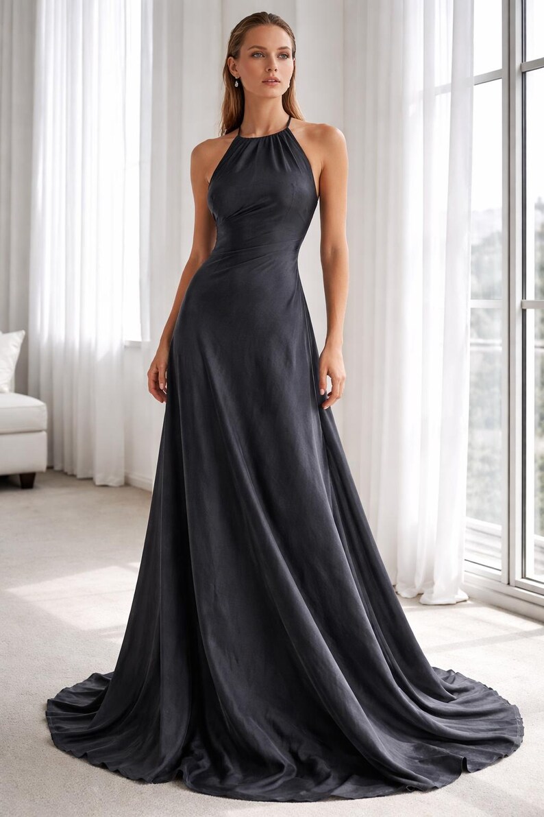 K&ouml;nnte beinhalten: Ein dunkelgraues Neckholder-Kleid mit flie&szlig;endem A-Linien-Rock. Das Kleid hat ein figurbetontes Oberteil und einen bodenlangen Saum. Das Model steht vor einem Fenster mit wei&szlig;en Vorh&auml;ngen.