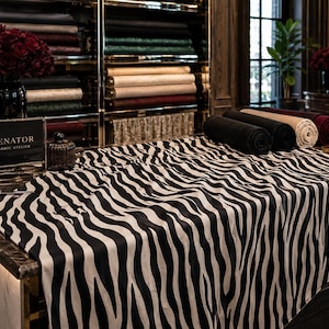 May include: A display of fabrics in a high-end atelier. A large zebra-striped fabric is draped over a counter. Rolled fabrics in black, brown, and burgundy are on the counter. Shelves in the background hold more fabric rolls.