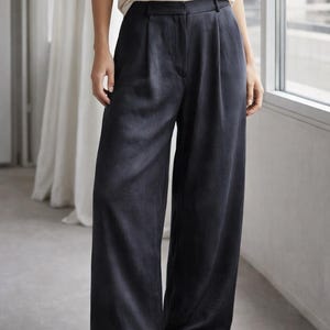 K&ouml;nnte beinhalten: Dunkelblaue Hose mit weitem Bein, hoher Taille und Falten. Die Hose wird mit einem wei&szlig;en &auml;rmellosen Oberteil und schwarzen Ballerinas kombiniert. Das Outfit ist schlicht und elegant.