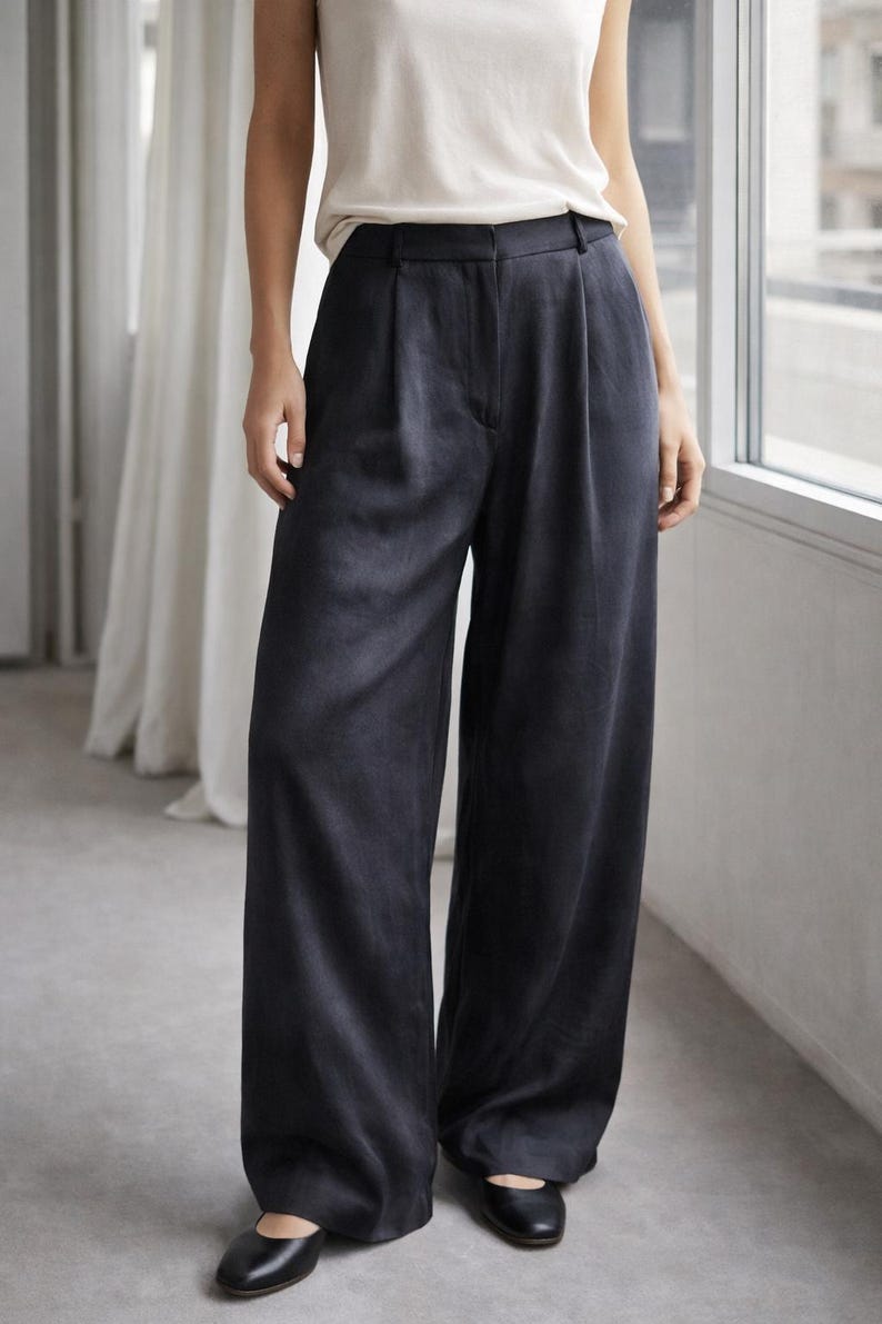 K&ouml;nnte beinhalten: Dunkelblaue Hose mit weitem Bein, hoher Taille und Falten. Die Hose wird mit einem wei&szlig;en &auml;rmellosen Oberteil und schwarzen Ballerinas kombiniert. Das Outfit ist schlicht und elegant.