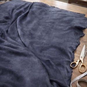 K&ouml;nnte beinhalten: Ein gro&szlig;es St&uuml;ck dunkelblauen Stoffes mit rohem Rand, drapiert auf einem Holztisch. Eine goldfarbene Schere und ein Ma&szlig;band liegen auf dem Tisch. Der Stoff scheint Leinen oder ein &auml;hnliches Material zu sein.