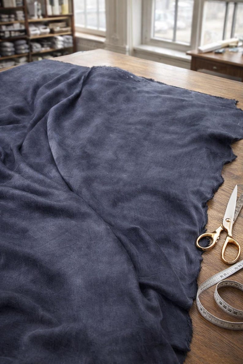 K&ouml;nnte beinhalten: Ein gro&szlig;es St&uuml;ck dunkelblauen Stoffes mit rohem Rand, drapiert auf einem Holztisch. Eine goldfarbene Schere und ein Ma&szlig;band liegen auf dem Tisch. Der Stoff scheint Leinen oder ein &auml;hnliches Material zu sein.