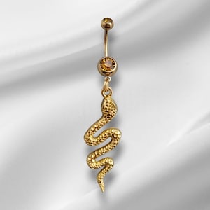 Può includere: Anello per ombelico color oro con un ciondolo a forma di serpente pendente. L'anello presenta una piccola gemma rotonda nella parte superiore e una gemma più grande al centro. Il ciondolo a forma di serpente è dettagliato con squame strutturate.
