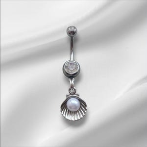 May include: A silver belly button ring with a dangling seashell charm. The ring features a clear gem and a pearl within the shell. The jewelry is displayed against a white background.
