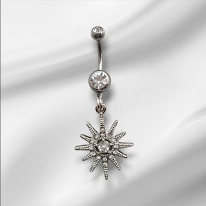 May include: A silver-colored belly button ring with a dangling starburst charm. The ring features a curved barbell design with a clear gem at the top and bottom. The starburst charm is detailed with multiple small clear gems.
