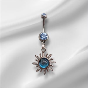 Puede incluir: Un piercing de ombligo plateado con un colgante de sol. El anillo presenta una gema azul claro en la parte superior y una gema azul más grande en el centro del sol. El colgante de sol tiene rayos detallados.