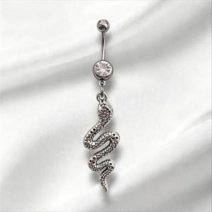 May include: A silver-toned belly button ring featuring a curved barbell with a clear round gemstone at the base. A dangling snake charm with detailed scale textures hangs from the bottom of the gemstone setting, creating a vertical design.