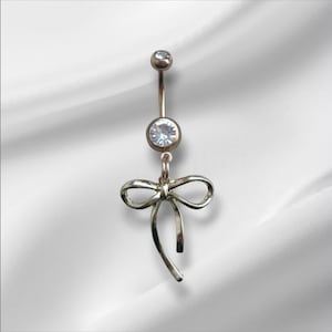May include: A gold-colored belly button ring with a bow charm. The ring features a small, clear gem at the top and a larger gem in the center. The bow charm hangs below the center gem.