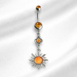 Puede incluir: Un piercing para el ombligo plateado con piedras preciosas de color ámbar y un colgante de sol. La barra curva tiene una pequeña gema en la parte superior y una más grande en el medio. El colgante de sol tiene una gema central.