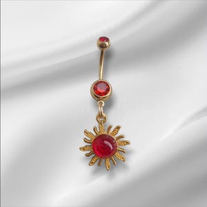 May include: Gold-coloured belly button ring featuring three red gemstones. The dangling charm is a sunburst design with a large, round red gem in the centre. The jewellery is displayed against a white background.