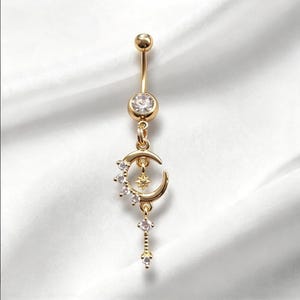 May include: A gold-toned navel ring featuring a clear round gemstone at the top, a crescent moon charm adorned with small clear stones, a central star detail, and a dangling vertical chain with two additional clear stones at the base.