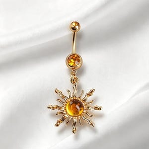 May include: A gold-toned navel barbell featuring a curved post with a small ball at the top and a larger circular setting containing a yellow gemstone. A sun-shaped charm with wavy rays and a central yellow gemstone hangs from the lower setting.