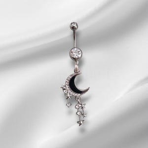 Puede incluir: Un piercing para el ombligo plateado con un diseño de media luna con esmalte negro y estrellas colgantes. El anillo tiene una gema transparente en la parte superior y una gema más grande debajo. Las estrellas y la luna están adornadas con pequeñas gemas transparentes.