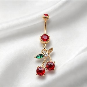 May include: A gold-tone navel ring featuring a curved barbell with a red crystal top ball and a larger red crystal bottom ball. A dangling cherry charm hangs from the bottom, accented with two red crystal cherries, a green crystal leaf, and a clear crystal.
