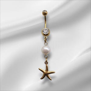 May include: Gold-coloured belly button ring with a dangling pearl and starfish charm. The ring features a clear gem and a small gold ball at the top. The starfish charm adds a beachy vibe to this body jewellery.