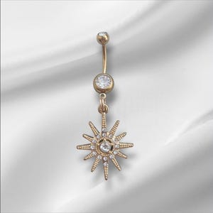May include: A gold-colored belly button ring with a sunburst charm. The ring features a curved barbell design with a small, round gem at the top and a larger gem in the center. The sunburst charm is adorned with multiple small gems.