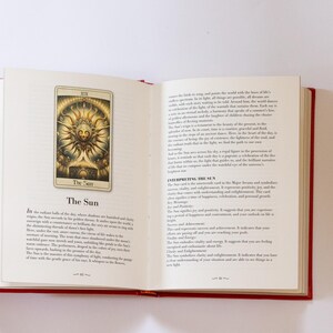 May include: An open book with red covers, displaying a tarot card illustration of "The Sun." The card features a golden sun with a face, surrounded by rays. Text on the page describes the card's meaning. The book is on a white surface.