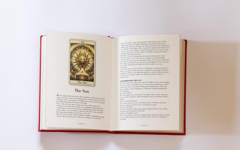 May include: An open book with red covers, displaying a tarot card illustration of "The Sun." The card features a golden sun with a face, surrounded by rays. Text on the page describes the card's meaning. The book is on a white surface.