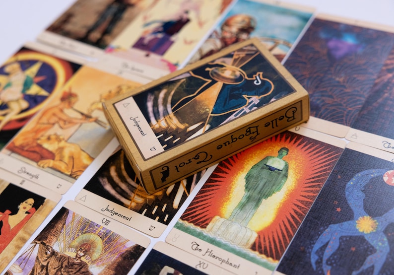 May include: A deck of Belle Epoque Tarot cards with a tan box featuring the word "Judgement". The cards have colorful illustrations with symbols and figures. The cards are spread out on a white surface.