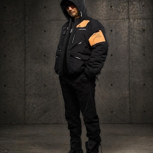 May include: Black hooded jacket with orange accents on the shoulders and upper arm. The jacket has multiple pockets and zippers. The person is wearing black pants and black boots. The background is a textured gray wall.