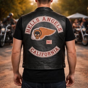 Puede incluir: Chaleco de cuero negro con el logo "Hells Angels" en la espalda, con una calavera alada y el texto "California". El chaleco se lleva sobre una camiseta negra y vaqueros azules. Se ven motocicletas al fondo.