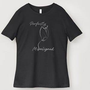 May include: A black short-sleeved t-shirt featuring a white line art illustration of a human silhouette. The text "Perfectly" is printed above the graphic, and the word "Misaligned" is printed below the graphic in a white cursive script font.