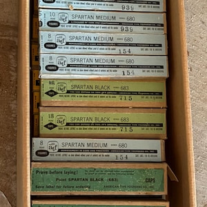 May include: A cardboard box filled with multiple boxes of Spartan Medium and Spartan Black paint. The boxes are green, white, and yellow with black text. The boxes are stacked inside the cardboard box.