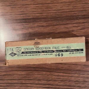 May include: A vintage rectangular box with the text "CENTURY SCHOOLBOOK ITALIC" and "465" printed on a green label. The box is brown and has the text "AMERICAN TYPE FOUNDERS CO., INC." and "069" printed on it. The box is on a wooden surface.