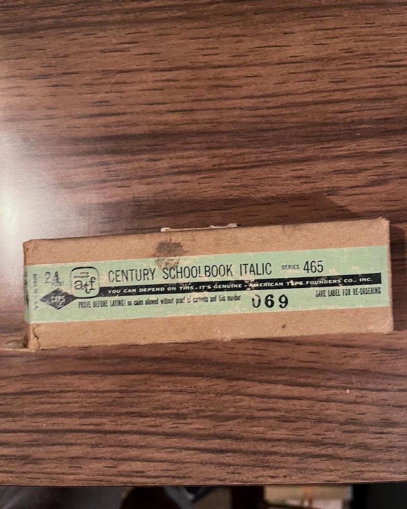 May include: A vintage rectangular box with the text "CENTURY SCHOOLBOOK ITALIC" and "465" printed on a green label. The box is brown and has the text "AMERICAN TYPE FOUNDERS CO., INC." and "069" printed on it. The box is on a wooden surface.
