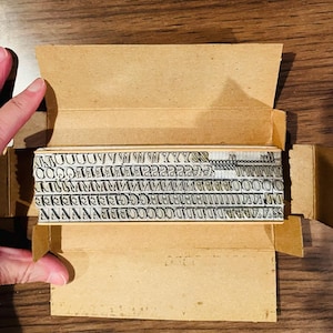 May include: A vintage metal letterpress type set in a cardboard box. The set includes various capital and lowercase letters, numbers, and symbols. The box is open, revealing the neatly arranged metal type blocks. The background is a wooden surface.