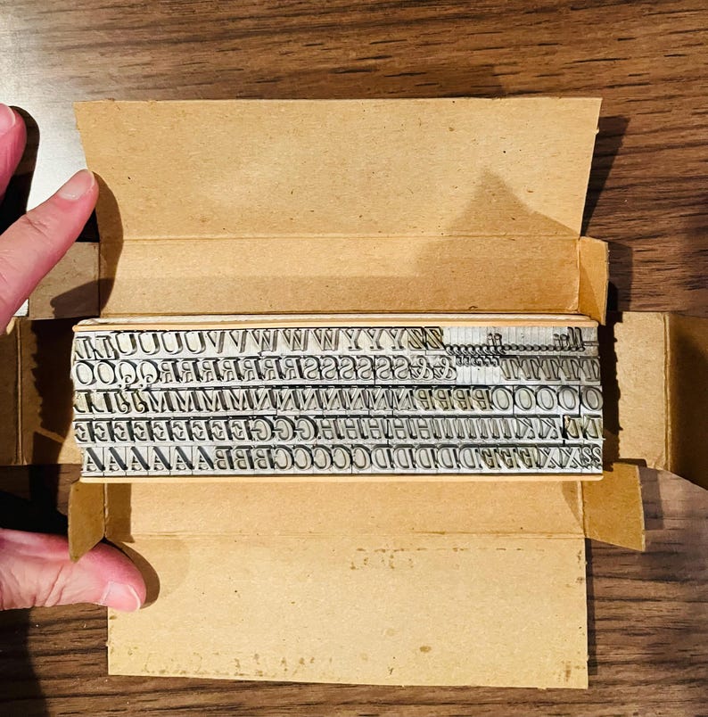 May include: A vintage metal letterpress type set in a cardboard box. The set includes various capital and lowercase letters, numbers, and symbols. The box is open, revealing the neatly arranged metal type blocks. The background is a wooden surface.