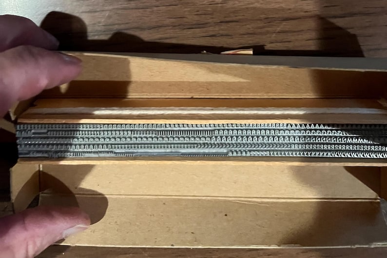 May include: A vintage wooden box containing metal letterpress type. The type is arranged in rows, with various fonts and sizes visible. The box is open, revealing the contents. The wood has a light brown color.