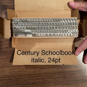 May include: A set of vintage metal letterpress type in a cardboard box. The type is arranged in rows and includes various letters and symbols. The box has the text "Century Schoolbook italic, 24pt".