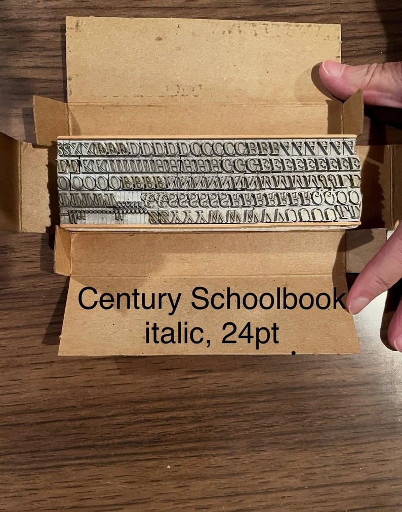 May include: A set of vintage metal letterpress type in a cardboard box. The type is arranged in rows and includes various letters and symbols. The box has the text "Century Schoolbook italic, 24pt".