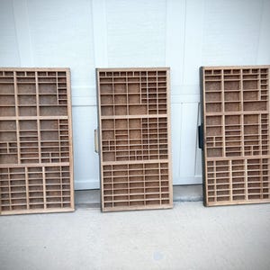 May include: Three wooden printer's trays with various sized compartments. The trays are made of light-colored wood with dark brown frames and handles. The trays are standing upright against a white wall.