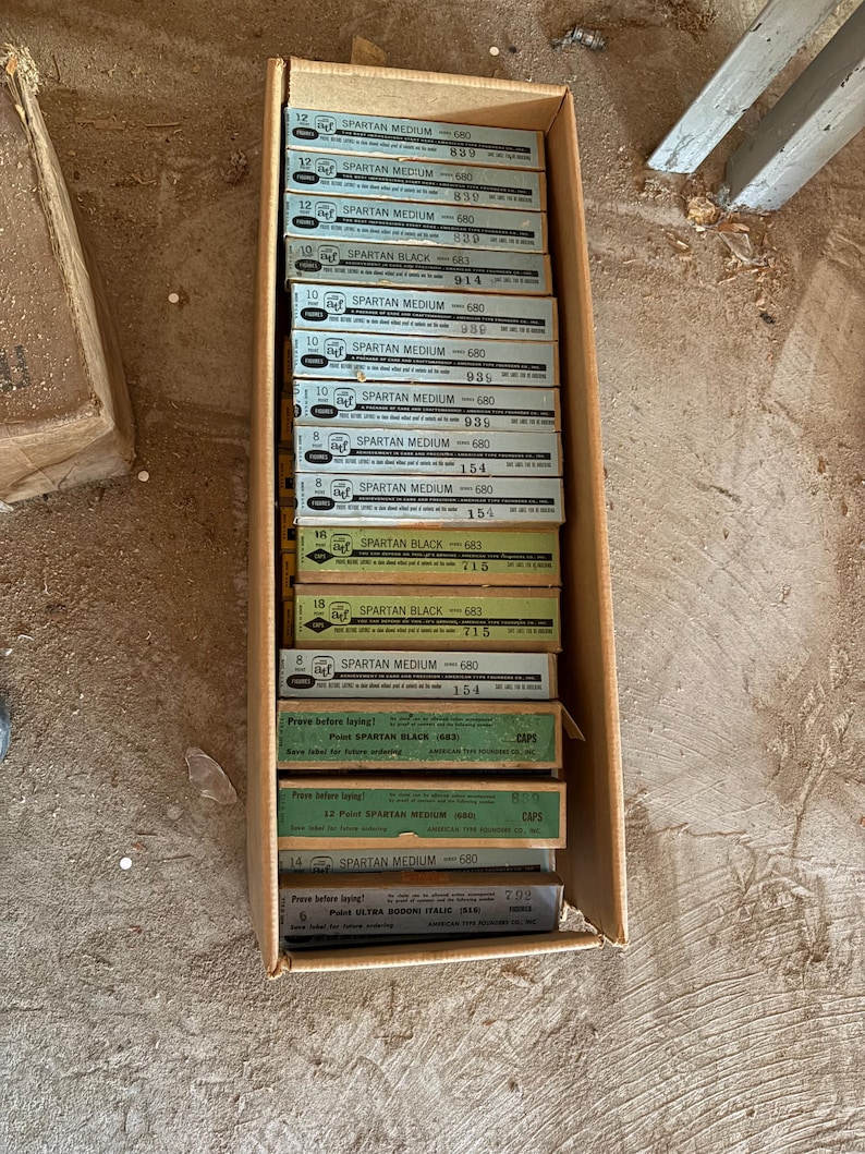 May include: A cardboard box filled with multiple boxes of Spartan Medium and Spartan Black paint. The boxes are green, white, and yellow with black text. The boxes are stacked inside the cardboard box.
