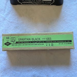 May include: A vintage Griswold Film Splicer and a box of Spartan Black caps. The film splicer is black with white text, and the box is green and tan with black text. The box reads "SPARTAN BLACK" and "AMERICAN TYPE FOUNDERS CO., INC."