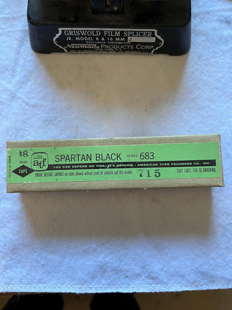 May include: A vintage Griswold Film Splicer and a box of Spartan Black caps. The film splicer is black with white text, and the box is green and tan with black text. The box reads "SPARTAN BLACK" and "AMERICAN TYPE FOUNDERS CO., INC."