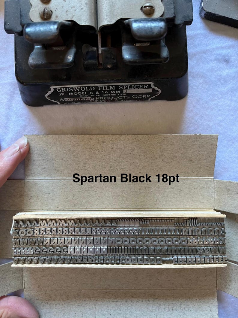 May include: A vintage Griswold Film Splicer, model 8 & 16 MM, with a metal construction. The image also shows a box of Spartan Black 18pt type, containing metal letter stamps. The text "Noumade Products Corp" is visible.