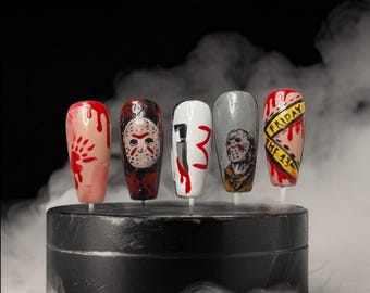 May include: Five press-on nails featuring horror-themed designs. One has red blood splatters, one a hockey mask, one a knife with the number 13, one a detailed character portrait, and one a yellow caution tape with the text FRIDAY THE 13TH.