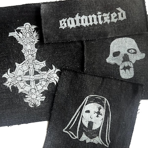 May include: Several black fabric patches with silver designs. The patches display a skull, a cross with skulls, a nun-like figure, and the word "satanized" in a gothic font. These patches are likely for clothing or accessories.