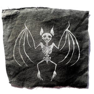 May include: A dark gray fabric patch featuring a white skeletal bat design. The bat has outstretched wings, a detailed rib cage, and a skull. The fabric has a slightly textured appearance, and the edges are frayed, giving it a handmade look.