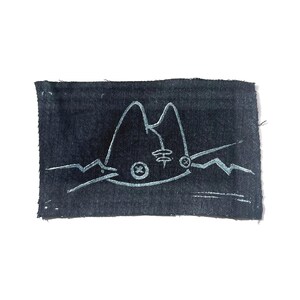 May include: A rectangular, dark blue fabric patch with a white, stylized cat face design. The cat's face features pointed ears, a circular eye, and a whisker on each side. The design is hand-drawn with a textured appearance.