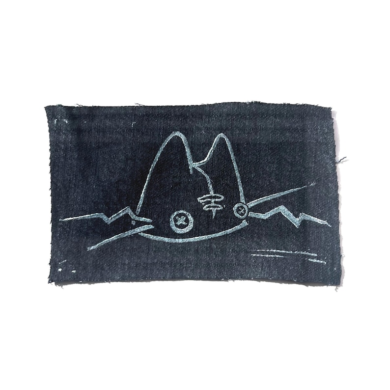 May include: A rectangular, dark blue fabric patch with a white, stylized cat face design. The cat's face features pointed ears, a circular eye, and a whisker on each side. The design is hand-drawn with a textured appearance.