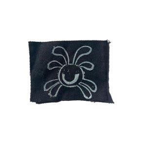 May include: A small, black fabric patch featuring a whimsical, smiling sun design in silver. The sun has curved lines for rays and a curved line for the mouth. The patch has a raw edge.